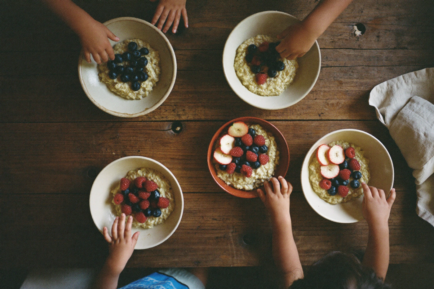 Kinderhände greifen an einem langen Holztisch nach Schüsseln mit Porridge und Beeren